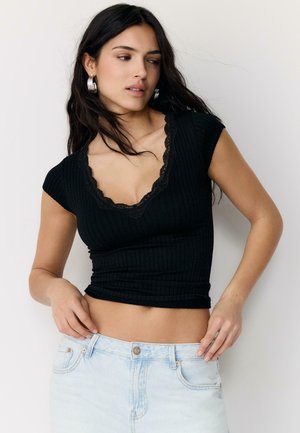 Kvinde med langt mørkt hår iført sølvfarvede creoløreringe, en sort ribbet crop top med blonder, og lyseblå jeans, poserer mod en ensfarvet baggrund.