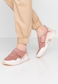 Sneakers w jasnoróżowym kolorze z dzianinową cholewką i syntetycznymi akcentami. Posiadają sznurowadła, amortyzowaną podeszwę oraz teksturowany wzór na materiale.