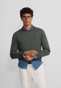 Suéter verde oliva de manga larga, con puños acanalados, cuello redondo, llevado sobre una camisa de denim clara, combinado con pantalones blancos.
