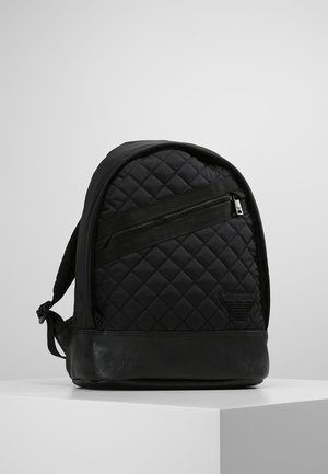 Black quilted backpack with diagonal front zipper, adjustable straps, and leather base standing on white pedestal against gray background.