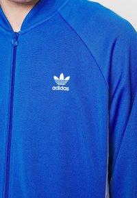 Sininen urheilullinen vetoketjullinen takki, valmistettu sileästä kankaasta, jossa valkoinen Adidas-logo ja kontrastivalkoiset yksityiskohdat vetoketjun ja hihojen varrella.