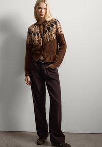Bruine gebreide cardigan met een crème en marineblauw patroon op de schouders, gecombineerd met donkere, wijde broekspijpen. De cardigan heeft een knoopsluiting en een geribbelde onderrand.