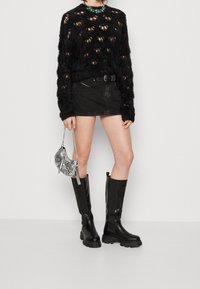Pull en tricot noir duveteux avec des gros trous, mini-jupe en jean noir, bottes noires à hauteur de genoux brillantes, et un sac à main argenté à motif serpent.