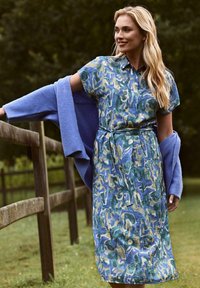 Knopenjurk in blauw, groen en tan abstract patroon; korte mouwen, tailleband, knie-lengte, gecombineerd met een lichtblauwe cardigan.