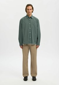 Jeune homme debout, portant une chemise verte ample à boutons, un pantalon beige et des chaussures marron foncé, sur un fond blanc uni.
