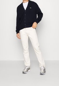Námořnický pletený cardigan s výstřihem do V a zapínáním na knoflíky, nošený přes bílý tričko, doplněný o krémové kalhoty a šedé sportovní tenisky.