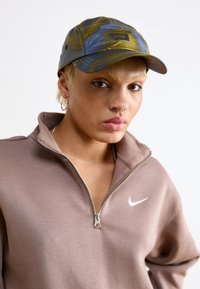 Cappellino blu e giallo con motivo, etichettato "RAINS", con visiera curva. Indossato con una felpa con zip color taupe caratterizzata da un logo Nike bianco.