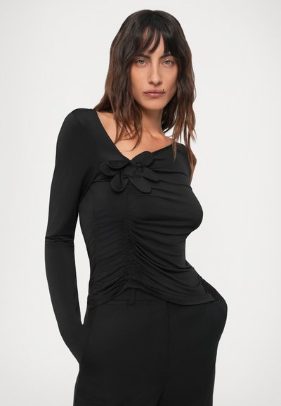 Hosszú, sötét hajú nő, aki ráncolt fekete hosszú ujjú felsőt visel, amelynek nyakánál egy nagy textilvirág részlet található, valamint fekete nadrágot.