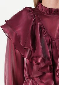 Bordeauxfarbene Bluse mit rüschierten Ärmeln und Ausschnitt; aus transparentem, glänzendem Stoff gefertigt; verfügt über geraffte Details und einen lockeren Schnitt.