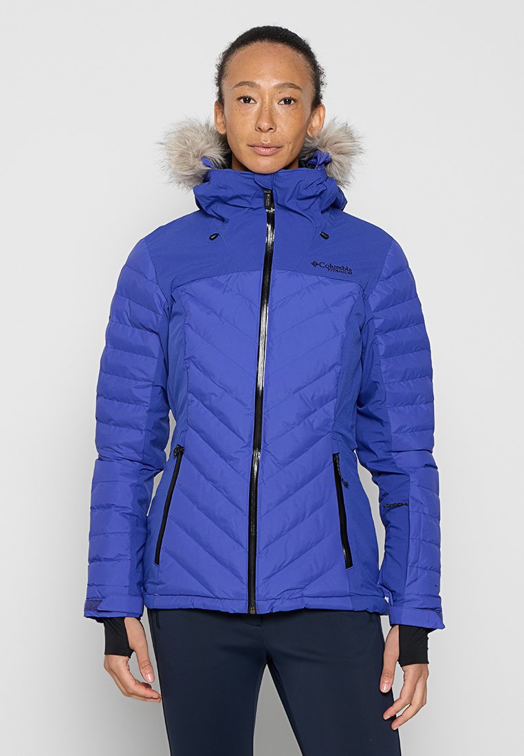 Veste matelassée bleue avec une capuche en fourrure, fermeture éclair à l'avant et motif en chevrons. Présente des zips noirs et des poignets élastiques pour un ajustement confortable.