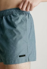 Costume da bagno in tessuto azzurro chiaro con un delicato motivo in rilievo di loghi. Presenta un elastico in vita e una piccola etichetta nera del marchio.