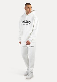 Sweatshirt à capuche blanc avec le texte noir "SMILODOX ATHL. WEAR", poche latérale, associé à un pantalon de sport assorti avec le logo sur la jambe.