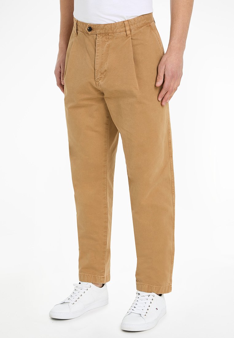 Pantaloni in cotone beige con vestibilità rilassata, caratterizzati da una piega frontale, passanti per cintura e chiusura con un solo bottone, abbinati a sneaker bianche.