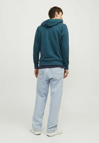 Sudadera teal para hombre con capucha ajustable, tejido suave y puños acanalados, combinada con unos vaqueros claros de corte holgado. Detalle de atuendo casual.