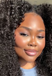 Nærbilde av en person med glansfulle, fyldige lepper, glatt hud, mørkt krøllete hår og naturlig sminke med en subtil høydepunkt på kinnbeina.