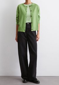 Cardigan verde lavorato a maglia con bottoni sopra una maglietta a righe bianche e verdi. Abbinato a pantaloni a gamba larga neri a vita alta e scarpe nere.