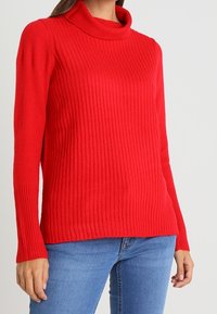 Punainen ribbikuvioitu korkeakauluksinen villapaita pitkillä hihoilla, jossa on teksturoidut neulekuviot ja väljä malli, yhdistettynä sinisiin farkkuihin.