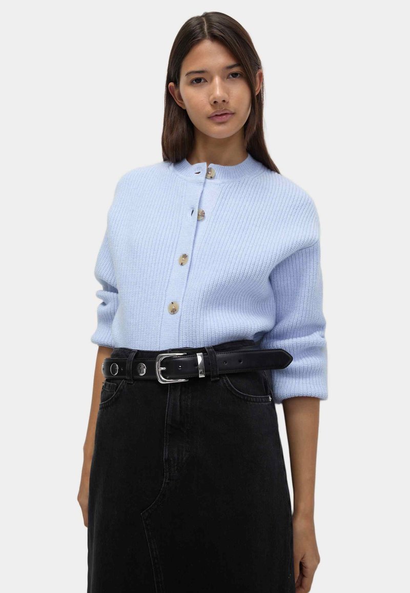 Cárdigan de punto acanalado de color azul claro con cuello redondo, con cuatro botones decorativos y llevado sobre una falda negra de talle alto con cinturón.