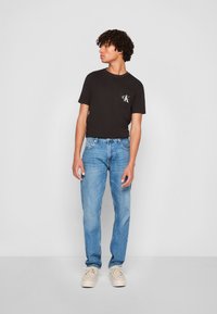 Zwart T-shirt met korte mouwen en een borstzakje met een wit logo, gecombineerd met lichtblauwe rechte jeans en beige schoenen.