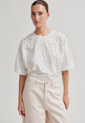 Young woman with pulled-back hair wears a white textured floral short-sleeve blouse tucked into high-waisted pale pants.
