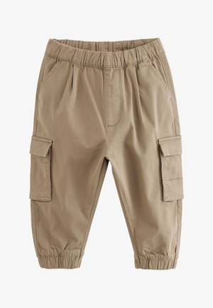 Småbarn med krøllet hår, der smiler, iført en mørk jakke, beige bukser og kondisko, siddende på en hvid overflade mod en neutral baggrund.