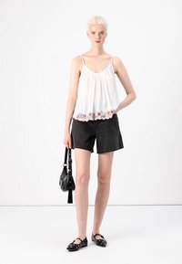 Biely bezrukávový top s vyšívaným kvetinovým lemom, kombinovaný s čiernymi džínovými šortkami, čiernou kabelkou a čiernymi slip-on topánkami. Hladká textura.