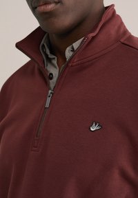 Vínový pulóver na zips s vysokým golierom, s čierno-bielym vyšívaným logom vtáka na ľavej strane hrudníka a sivou košeľou s golierom pod ním.