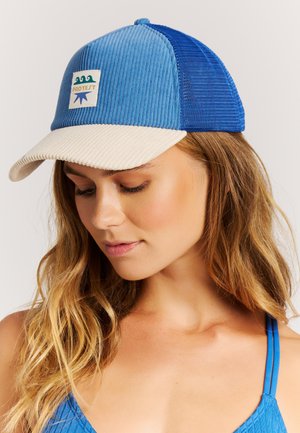 Vrouw met lang golvend haar, een blauw-beige corduroy pet en een blauw mouwloos topje, kijkt omlaag tegen een effen achtergrond.