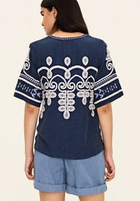 Mörkblå topp med vit intrikat broderi som har blommönster och swirl-mönster, rund halsringning och korta ärmar, ihop med ljusblå shorts.