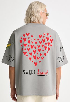 Persona che indossa una maglietta grigia con cuori rossi che formano una grande forma a cuore e le parole "SWEET heart" stampate sotto in nero e rosso.