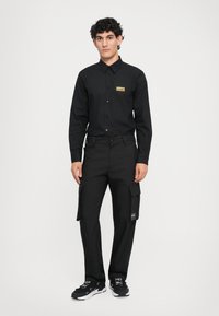 Svart skjorte med knapper og et gull logo-patch, sammen med svarte cargo-bukser med lommer, teksturert stoff og svarte sneakers.
