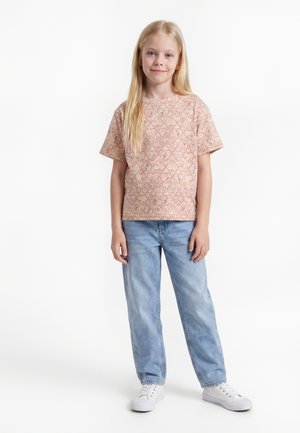 Ung pige med langt blondt hår iført en lyserød mønstret t-shirt, lyseblå jeans og hvide sneakers, der står og smiler.