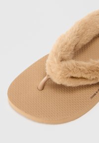 Tan flip-flop sandal med en teksturert gummisåle. Myke, fluffy stropper i beige, med en enkel gullaccent nær bunnen.