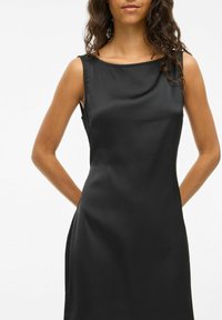 Femme aux cheveux ondulés portant une robe sans manches en satin noir, se tenant avec les bras derrière le dos contre un fond blanc uni.