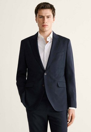 Young man wearing a dark blue suit and white striped dress shirt, standing with one hand in pocket against plain light background.