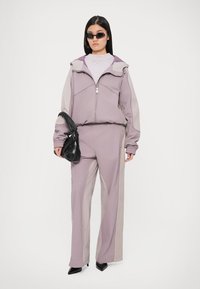 Δίπατο σκούφο σε ανοιχτό μωβ με αντίθετες μπεζ πάνελ. Περιλαμβάνει ζακέτα με φερμουάρ, ελαστικούς καρπούς και κομψά μαύρα αξεσουάρ.