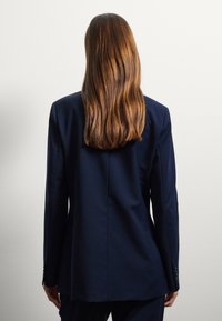 Donna con lunghi capelli castani che indossa un blazer blu scuro, in piedi di fronte a una semplice parete grigio chiaro.