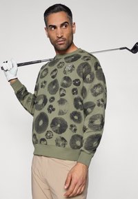 Πράσινο φούτερ με μαύρα κυκλικά και λουλουδένια σχέδια, με ριμπ μανσέτες και τελείωμα. Το μοντέλο κρατάει ένα ρόπαλο γκολφ φορώντας μπεζ παντελόνι.