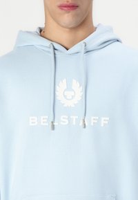 Világoskék kapucnis pulóver, elülső zsebbel, fehér logóval és "BE-STAFF" felirattal, ezüst végű húzózsinórokkal.