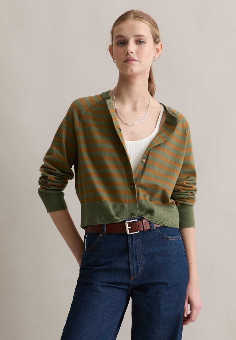 Vrouw draagt een groen-bruin gestreepte cardigan over een wit tanktopje, blauwe spijkerbroek en een bruine riem, staand tegen een effen beige achtergrond.