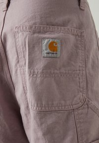 Lys lilla stoffshorts med en baklomme som har en firkantet, grå etikett med en oransje logo. Teksturert materiale med synlige søm detaljer.