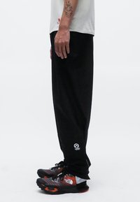Pantalons noirs en tissu doux, avec une coupe décontractée, des poches latérales et un petit logo sur le bas de la jambe gauche.