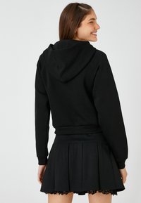 Sudadera negra con capucha y dobladillo acanalado, combinada con una falda plisada negra que presenta detalles de encaje. El modelo está de perfil.