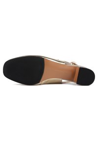 Chaussure à talon beige avec une bride transparente, présentant une tige lisse et une semelle en caoutchouc noir texturé pour plus de stabilité.