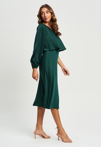 Zelena satenska midi obleka z večplastnim, drapiranim zgornjim delom in dolgimi napihnjenimi rokavi. Ima tekočo silhueto in je kombinirana z beige sandali z trakovi.