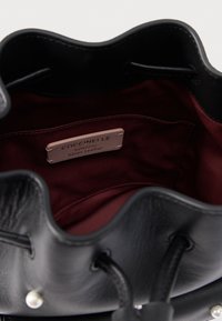 Binnenkant van een zwarte leren bucketbag met bordeauxrode voering en een label met de tekst "Coccinelle Authentic Italian Leather." Zachte textuur, afgeronde bovenkant.