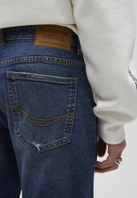 Jean en denim de teinte foncée avec une finition texturée. Comprend un patch de marque en cuir, des accents de surpiqûres jaunes et un design sur la poche arrière.