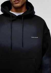 Donker hoodie met een verloop van zwart naar blauw, voorzien van koordjes, een voorkeurszak en de tekst "YOUNG ATELIER" in het wit.