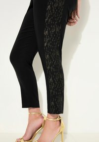 Des leggings noirs présentent un motif de rayure verticale dorée sur le côté. La personne porte des talons à brides dorés. Tissu lisse et ajusté.