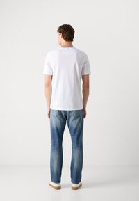 Hvid kortærmet t-shirt med et rektangulært patch på den øvre ryg, parret med lyseblå denimjeans med subtil falming og brune sko.